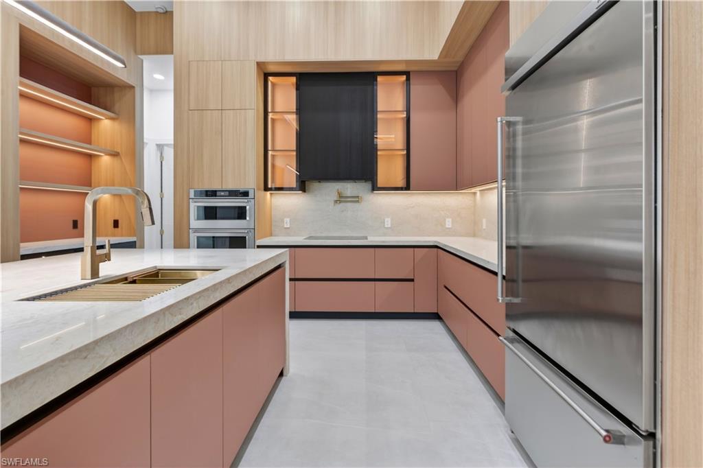Kitchen featuring a large center island with integrated sink and faucet, custom cabinetry with pink and wood-finish panels, stainless steel appliances including a refrigerator and double ovens, and recessed lighting