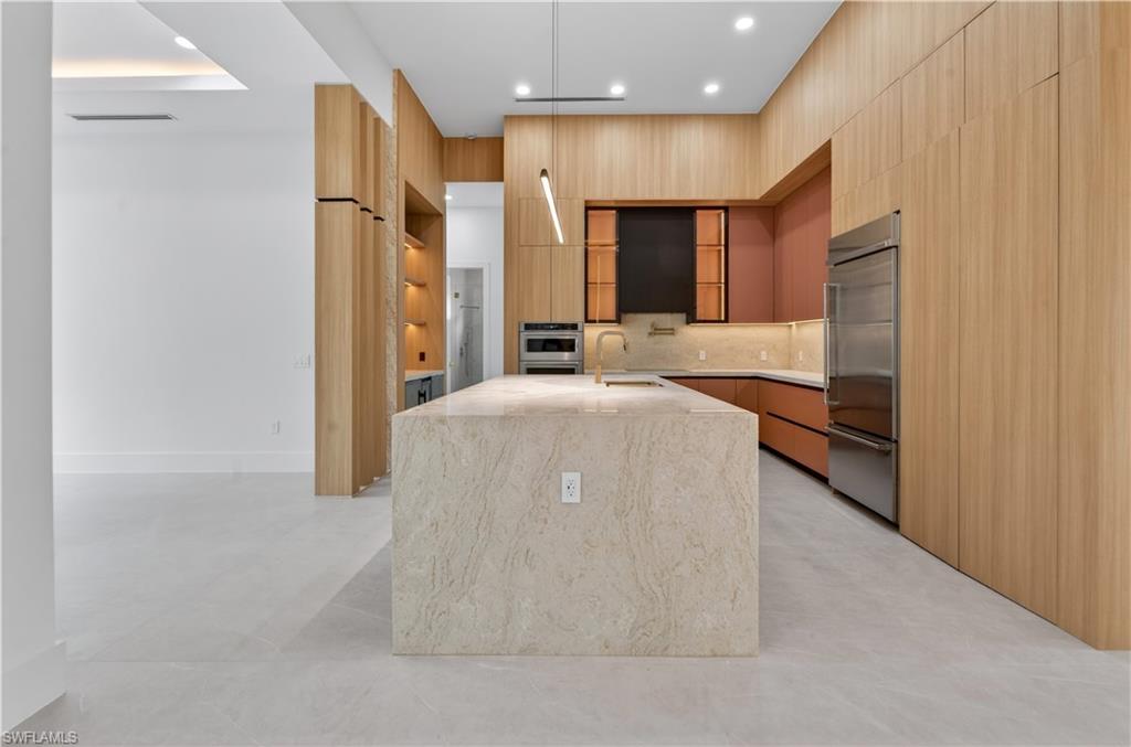 Modern kitchen featuring a large stone-finish island with integrated sink and gold-tone faucet, light wood-finish cabinetry, stainless steel refrigerator, and recessed lighting