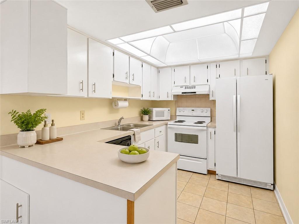 Bright kitchen featuring white cabinetry, light-toned countertops, a full suite of white appliances, a double basin stainless steel sink, and tile flooring