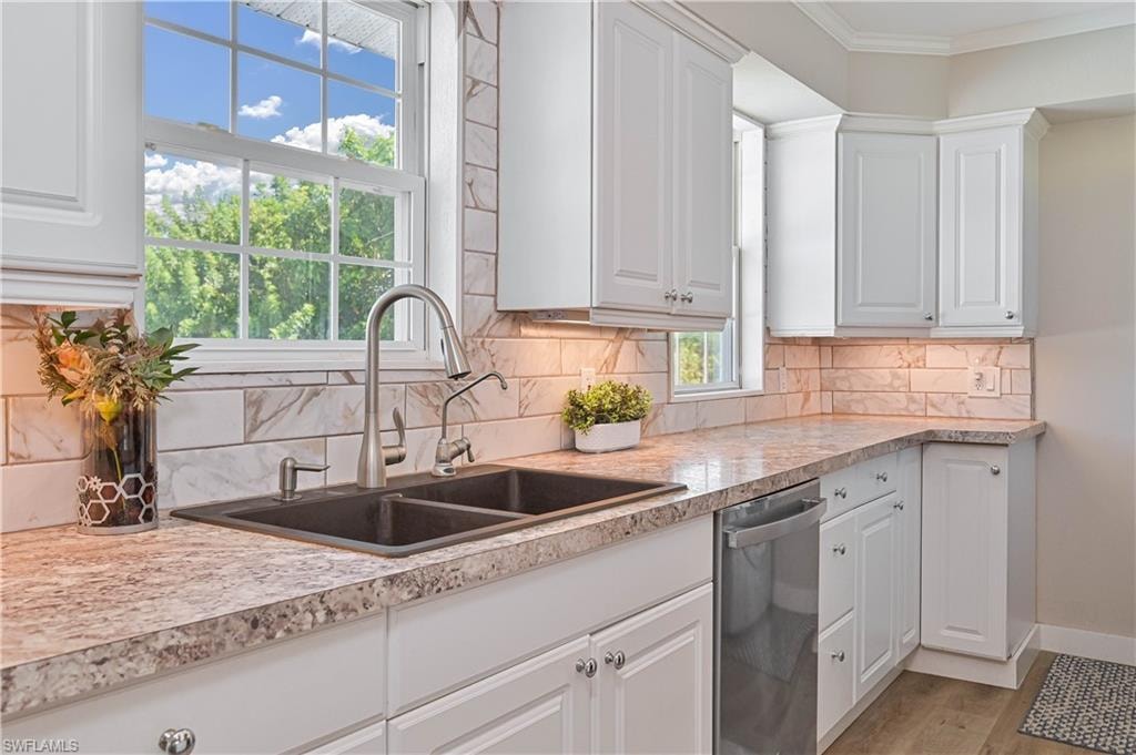 White cabinetry with crown molding, double basin sink with gooseneck faucet, light-colored countertops, tiled backsplash, and stainless steel dishwasher
