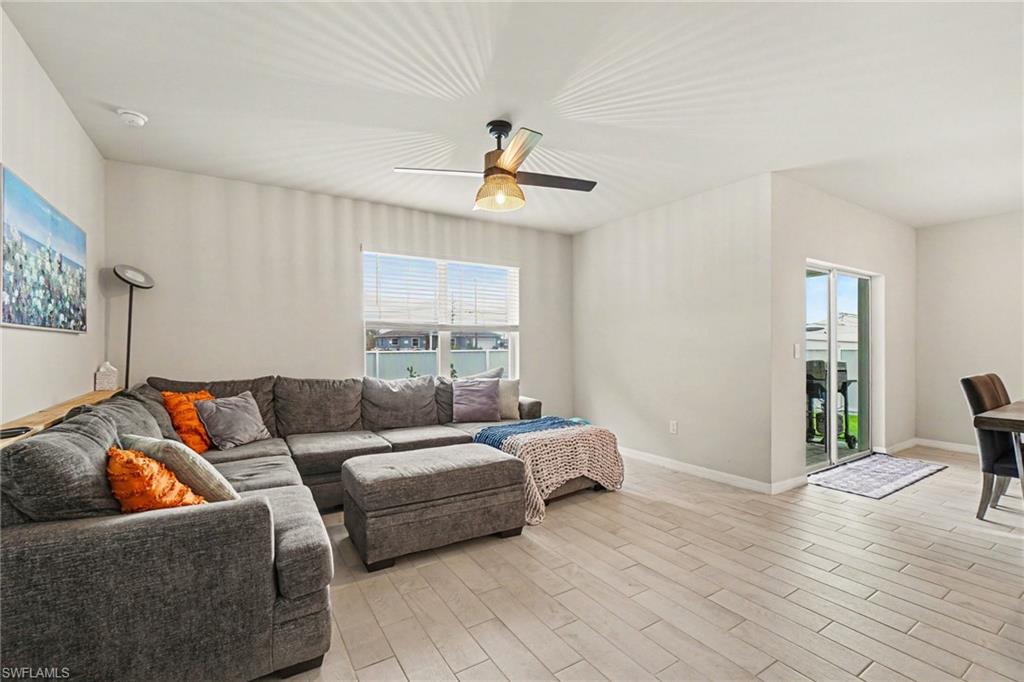 Spacious living area featuring light-toned walls, wood-finish flooring, a ceiling fan with integrated lighting, and a large window with horizontal blinds