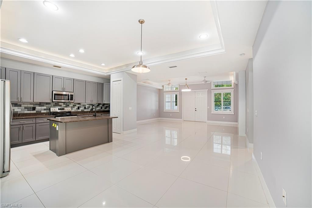 Kitchen featuring backsplash, appliances with stainless steel finishes, a raised ceiling, an island with sink, and pendant lighting