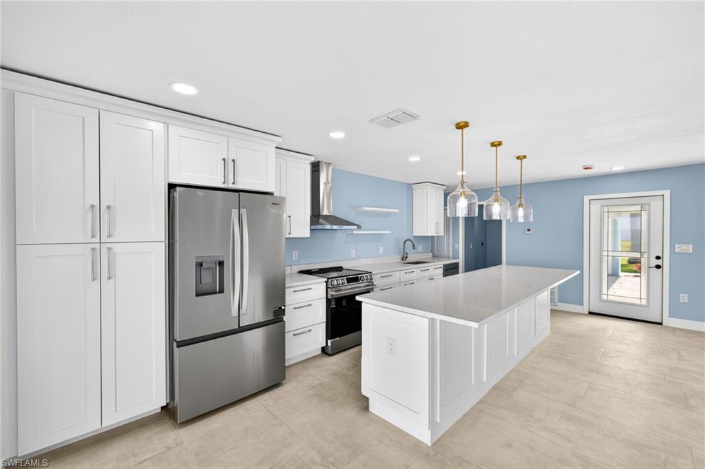 Modern kitchen featuring white shaker cabinetry, stainless steel appliances, a center island with a light-toned countertop, three glass pendant lights with brass accents, and light-toned tile flooring