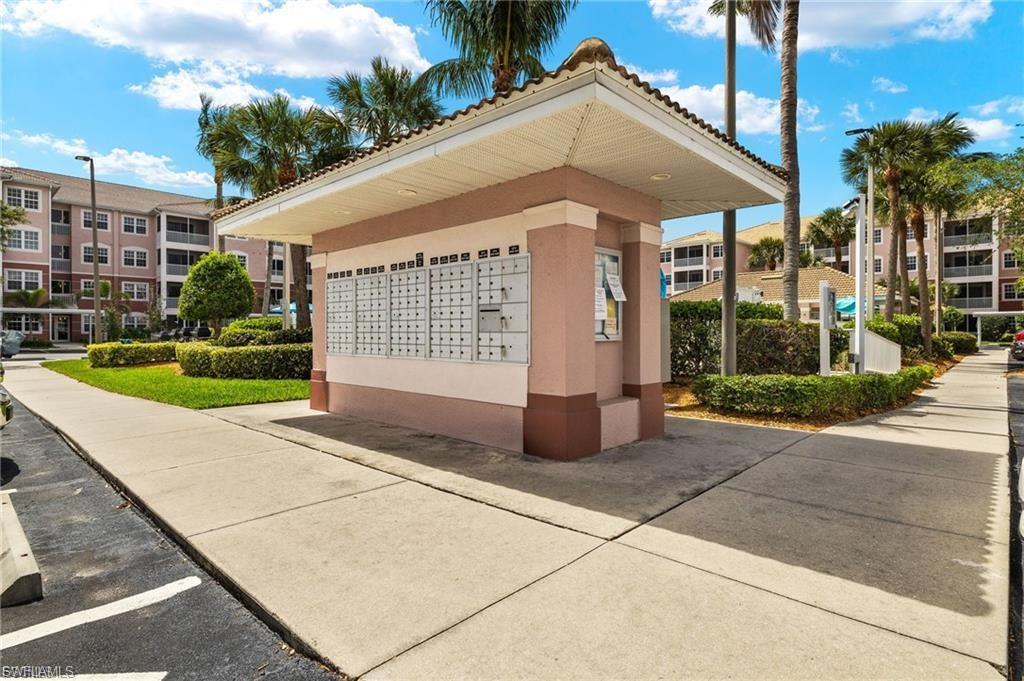 Community mailbox structure featuring multiple mail receptacles and a tiled roof, surrounded by manicured landscaping, paved walkways, and palm trees