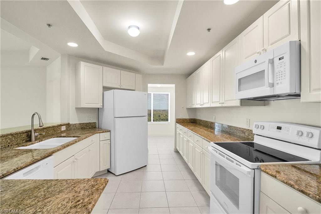 Kitchen featuring white appliances, a tray ceiling, recessed lighting, white cabinetry, and dark stone counters