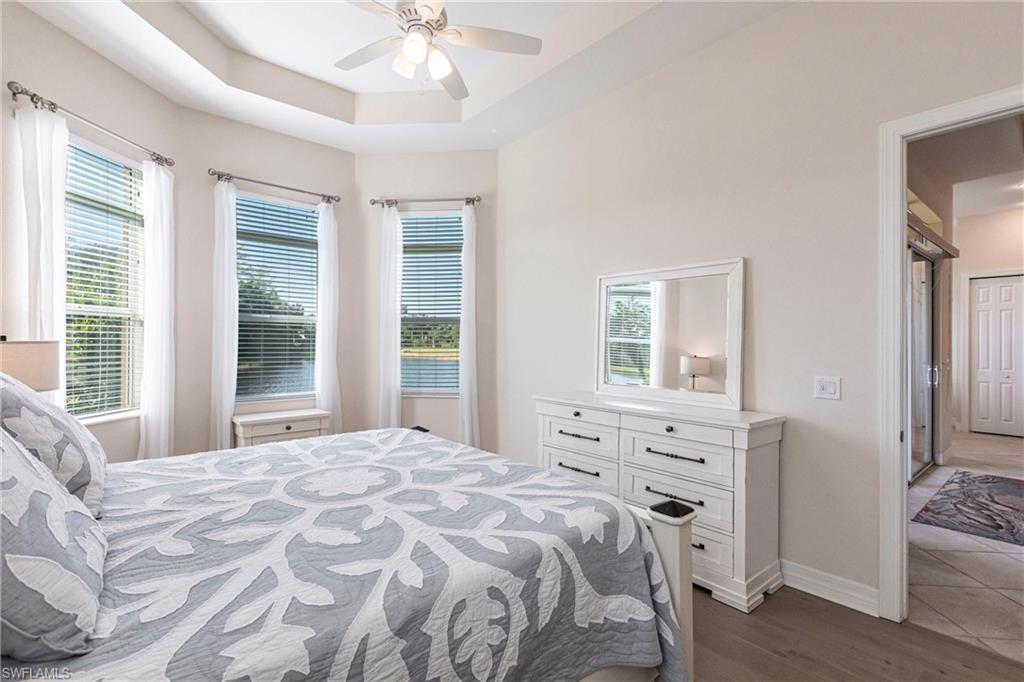 Bedroom featuring a tray ceiling with a ceiling fan, three windows with horizontal blinds and sheer curtains, wood-finish flooring, and a doorway opening to an adjacent hallway
