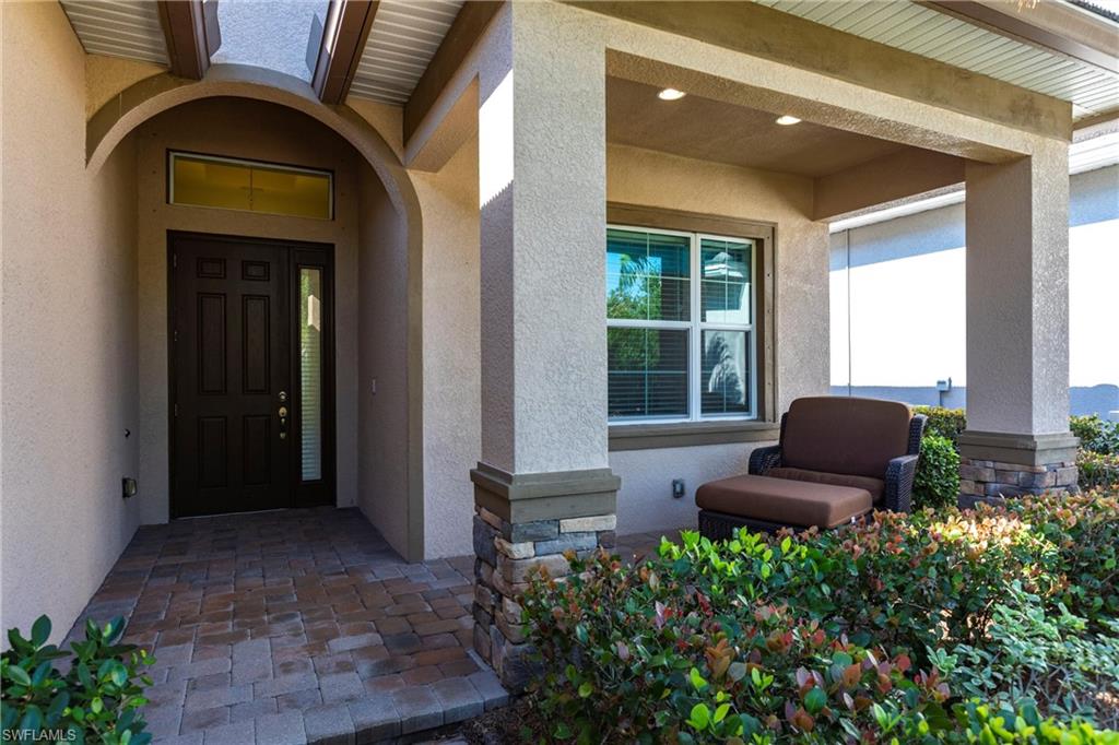 Inviting front entrance featuring a dark paneled door with a sidelight and transom window, a covered porch with recessed lighting, stone-clad pillars, and paver flooring