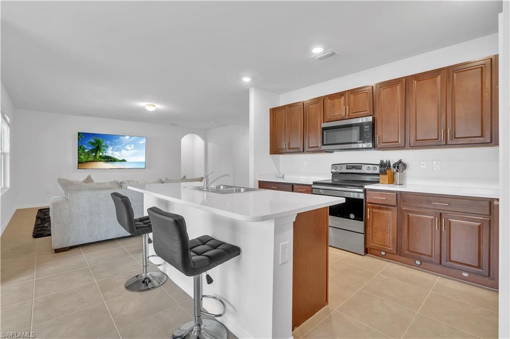 Kitchen featuring appliances with stainless steel finishes, arched walkways, a breakfast bar area, light tile patterned flooring, and brown cabinets