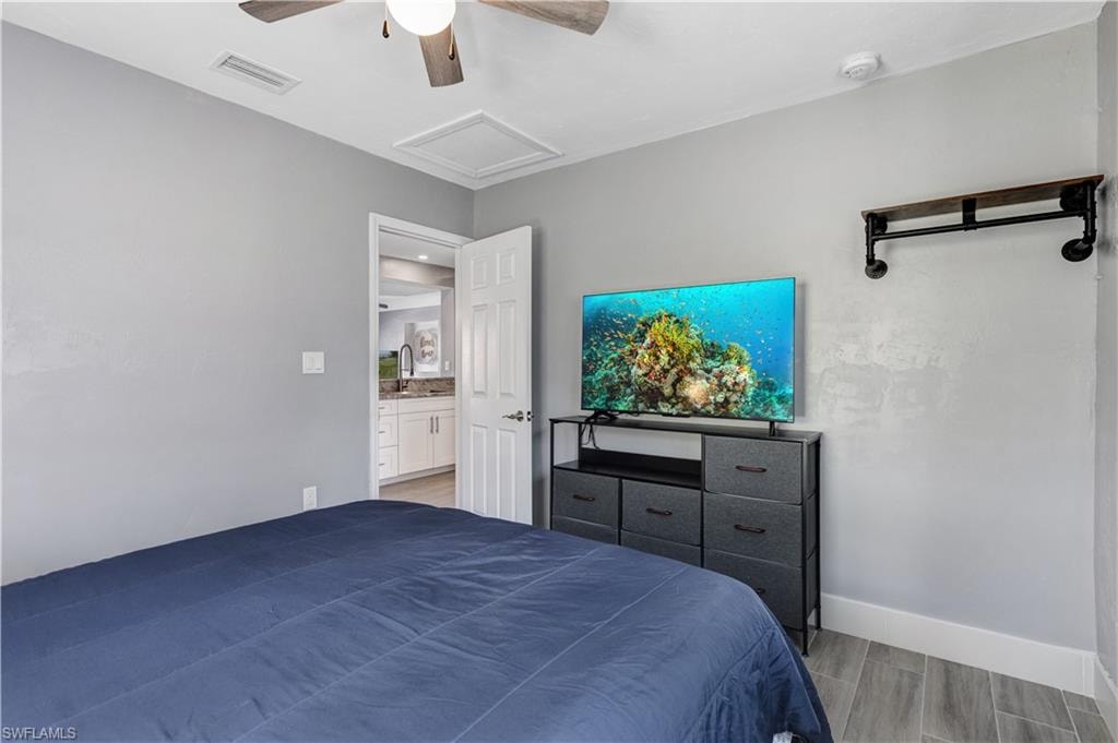 Bedroom featuring a ceiling fan, light gray wall finishes, wood-finish tile flooring, and a white six-panel door leading to a bathroom with a white vanity and dark faucet