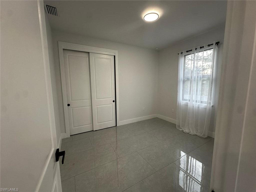 Room featuring light gray tile flooring, a window with sheer white curtains, a flush-mount ceiling light fixture, and a closet with white sliding doors and black hardware