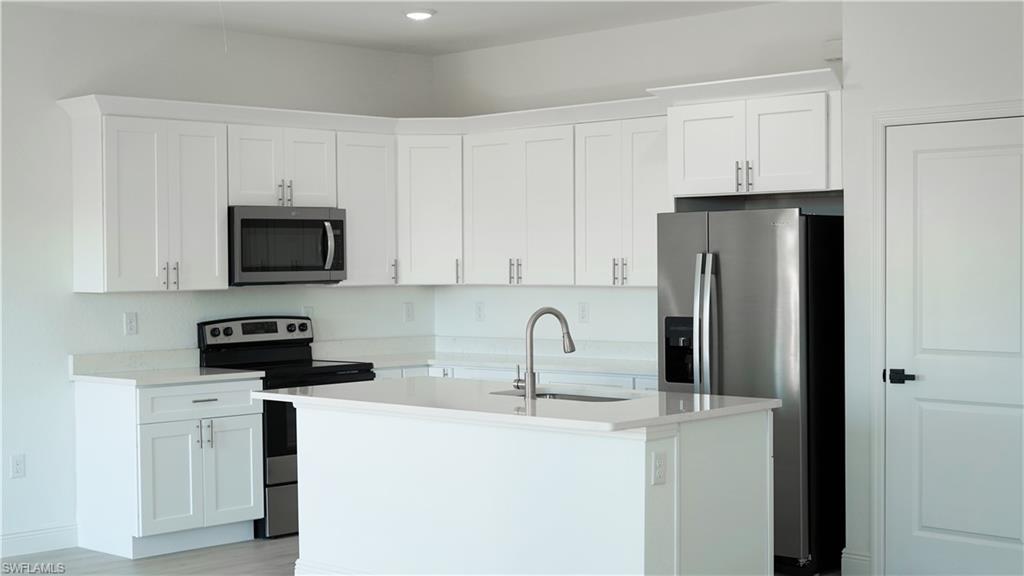 Modern kitchen featuring white shaker cabinetry, stainless steel appliances, a center island with sink and gooseneck faucet, light-toned countertops, and recessed lighting