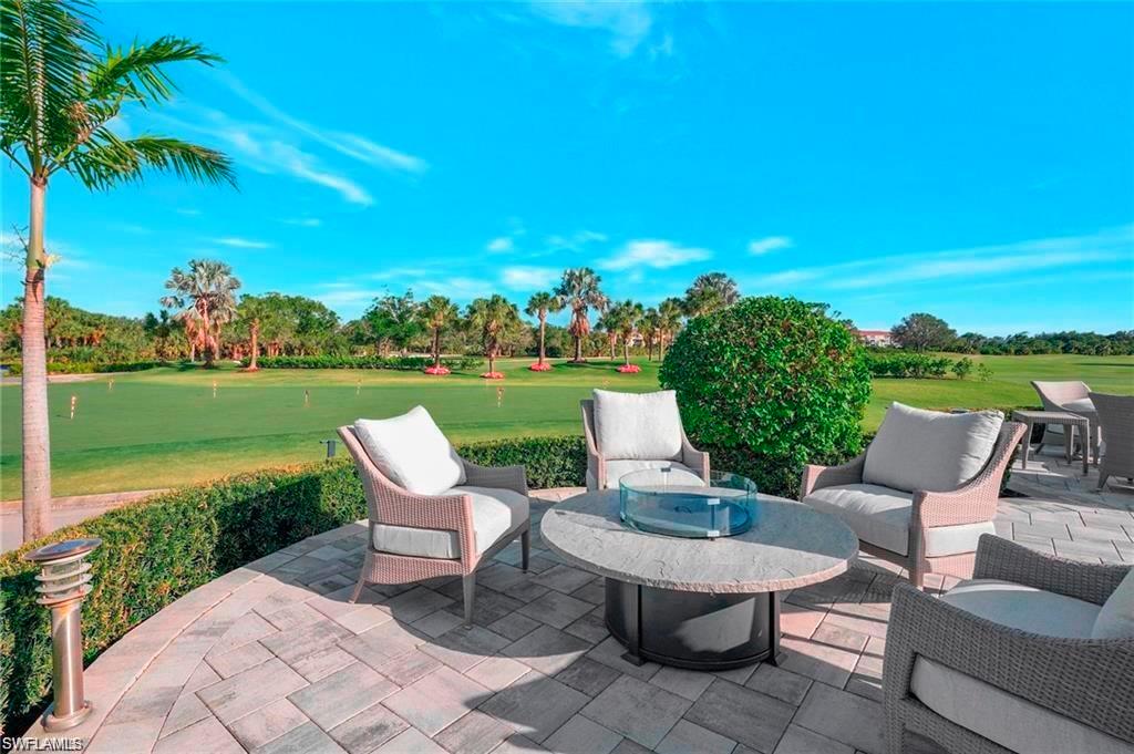 Paver patio with a fire pit table, surrounded by manicured hedges and a palm tree, overlooking a vast green expanse