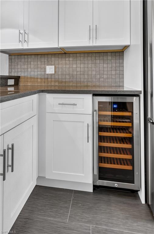 Stainless steel beverage cooler built into white shaker cabinetry with dark stone countertops, mosaic tile backsplash, and large format floor tiling