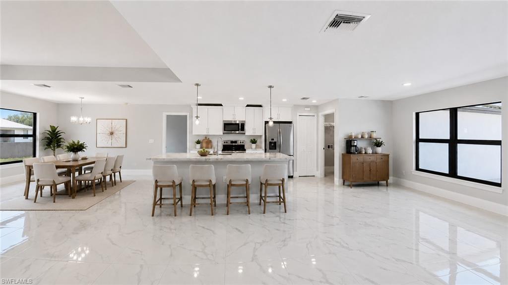 Expansive open-concept living area featuring white tile flooring, a large kitchen island with pendant lighting, white cabinetry, and stainless steel appliances