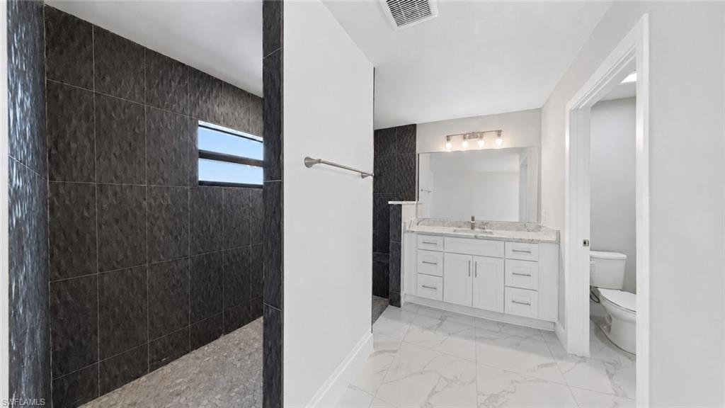 Modern bathroom featuring a large dark-tiled shower with a horizontal window, white vanity with dual sinks, spacious mirror, and elegant white marble-finish floor tiling