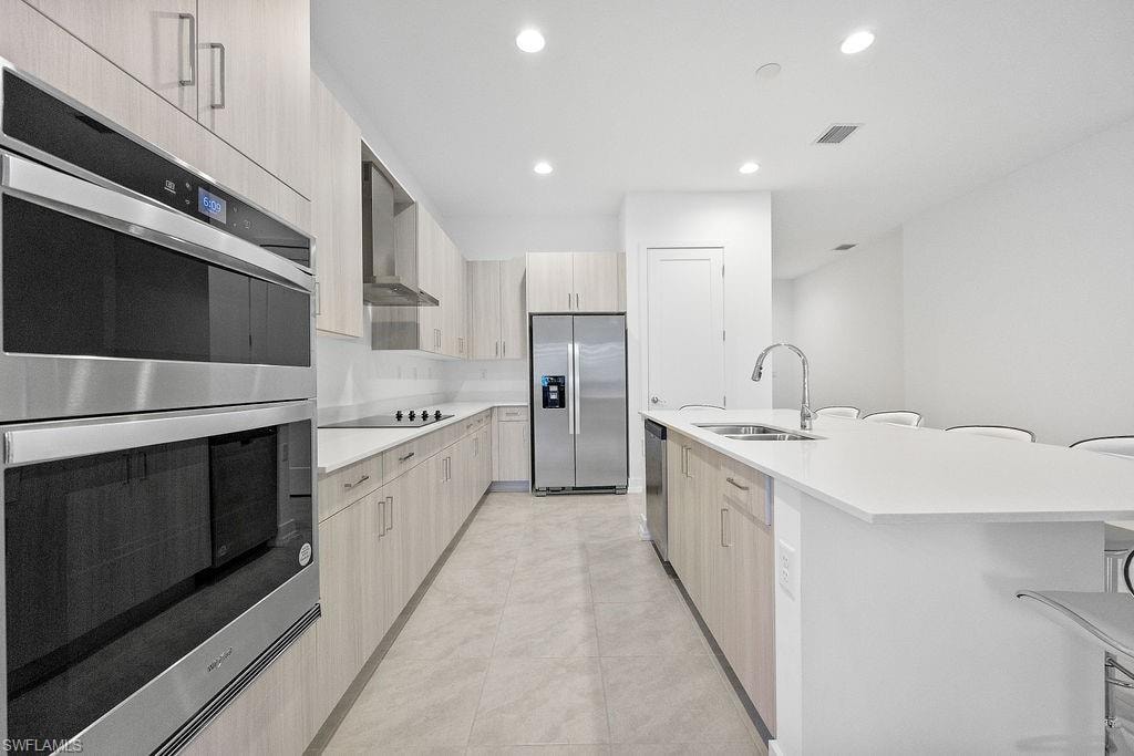 Kitchen with an island with sink, light tile flooring, wall chimney exhaust hood, sink, and appliances with stainless steel finishes