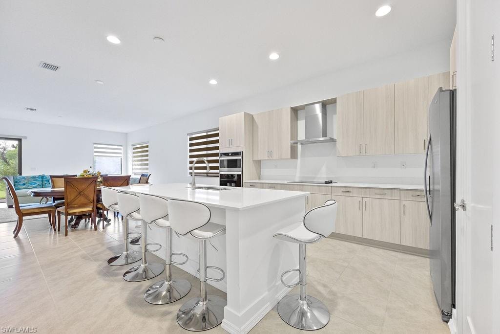 Kitchen with an island with sink, stainless steel appliances, light tile floors, wall chimney exhaust hood, and sink