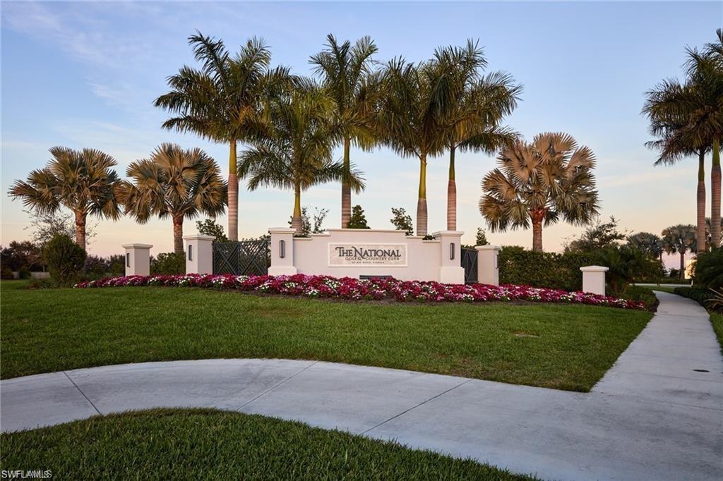 Property entrance featuring a substantial sign, lush green lawn, vibrant floral landscaping, mature palm trees, and a paved walkway