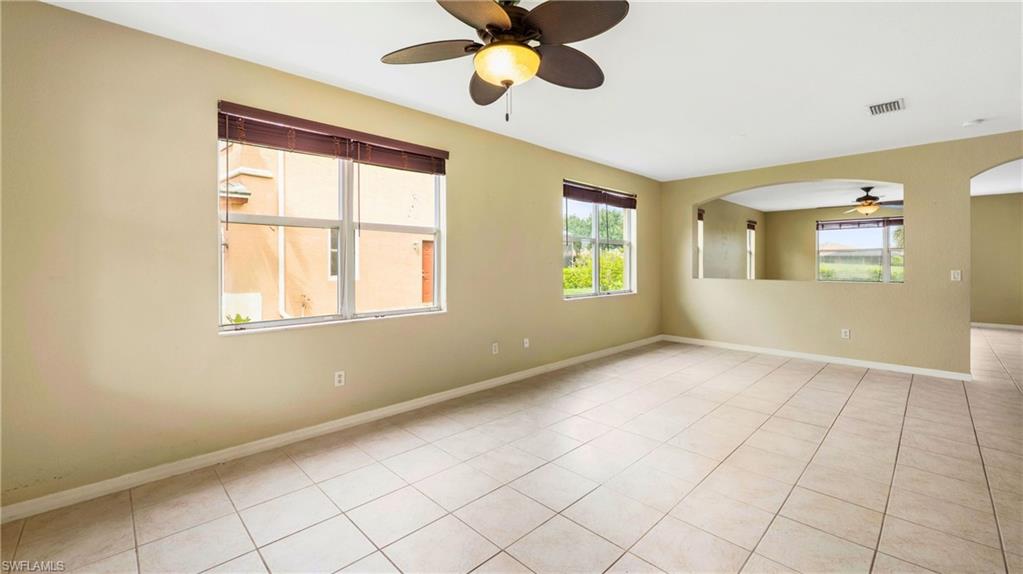 Spacious interior room featuring light-toned tile flooring, multiple windows with brown blinds, a ceiling fan with integrated lighting, and an arched half-wall opening