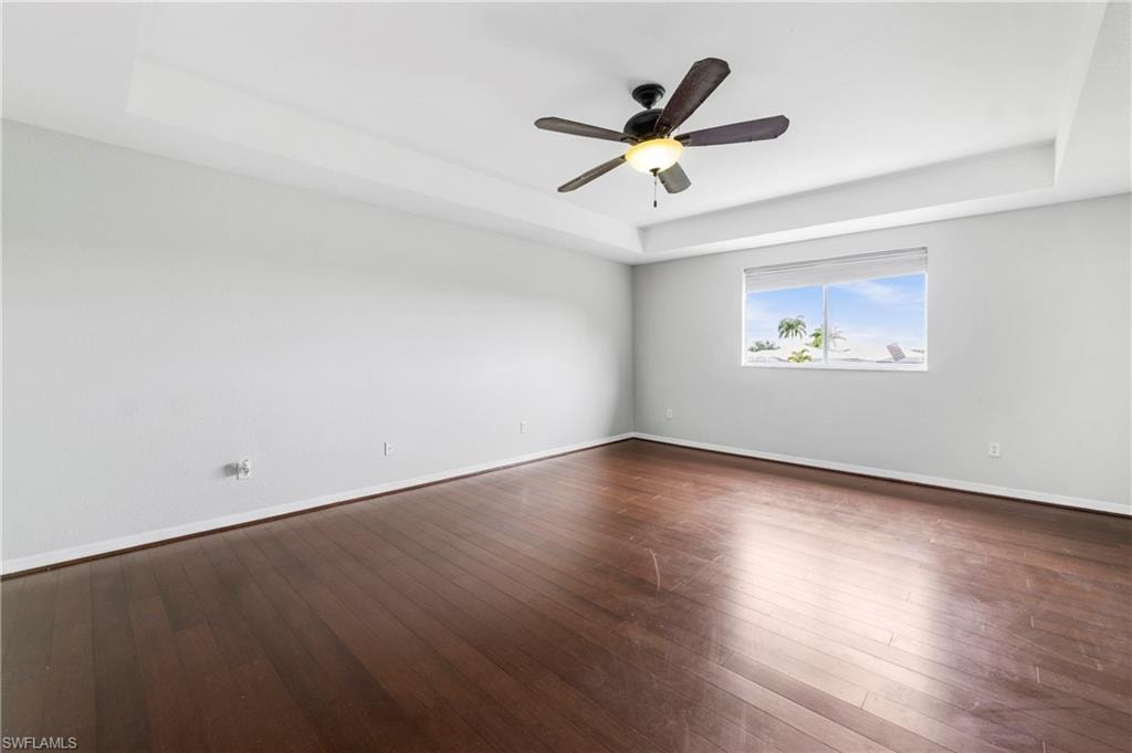 Spacious room featuring rich wood-finish flooring, a tray ceiling, and a ceiling fan with integrated lighting