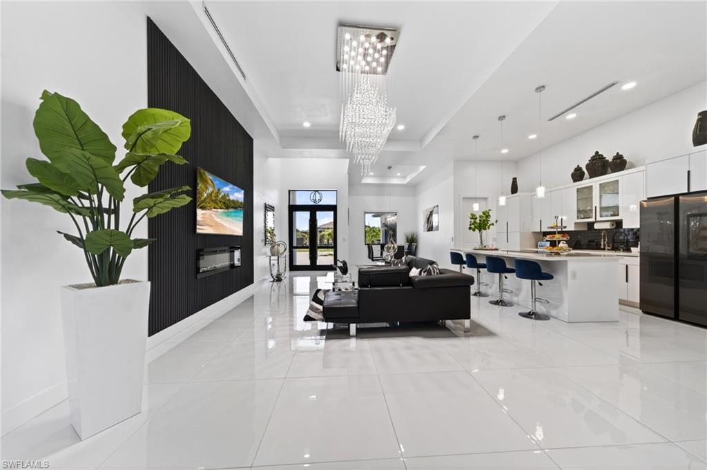 Spacious open-concept interior featuring large format white tile flooring, a contemporary linear fireplace, an elegant crystal chandelier, a kitchen island with seating, and white flat-panel cabinetry with glass accents