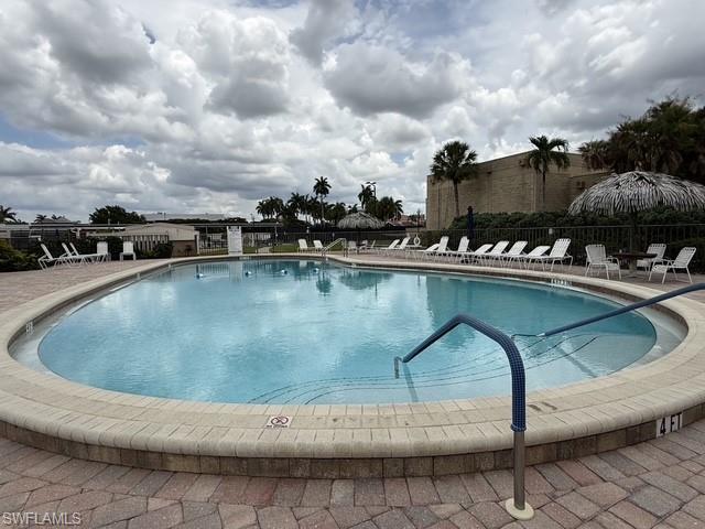 Curved swimming pool featuring a brick paver deck, a stucco building in the background, and surrounding palm trees