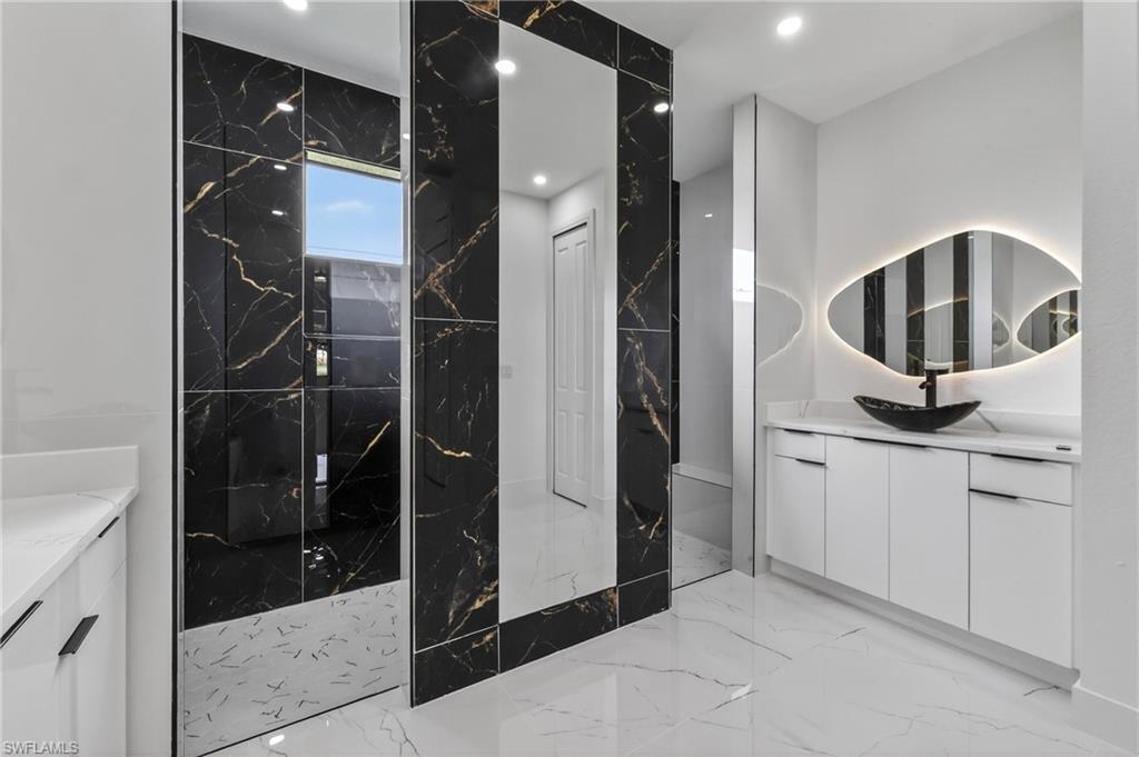 Contemporary bathroom featuring black and gold veined accent walls, white cabinetry with integrated pulls, a vessel sink, and an illuminated organic-shaped mirror