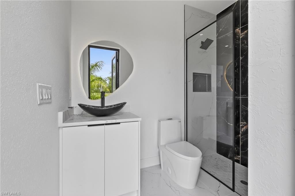 Modern bathroom featuring a glass-enclosed shower with black-veined wall tile, an irregular-shaped illuminated mirror, and a black vessel sink set on a white vanity with a light-colored countertop