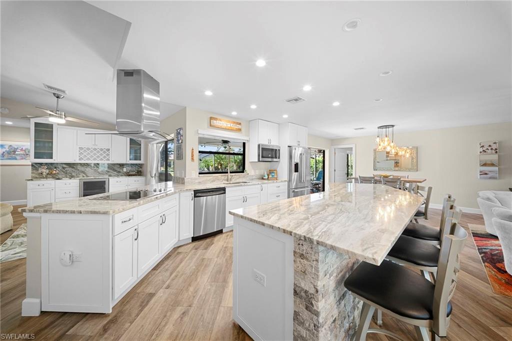 Spacious kitchen featuring an expansive island with stone veneer detailing, white shaker-style cabinetry, stainless steel appliances, and wood-finish flooring