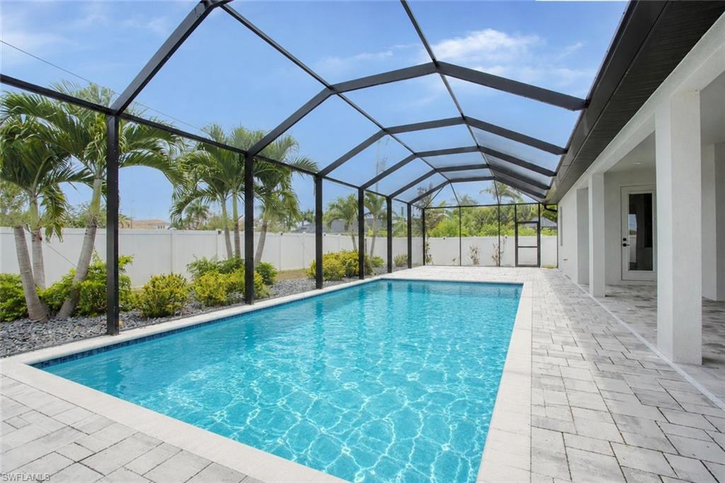 Screened-in pool area featuring a rectangular pool with blue tile accents, light gray paver decking, a white privacy fence, mature palm trees, and lush landscaping