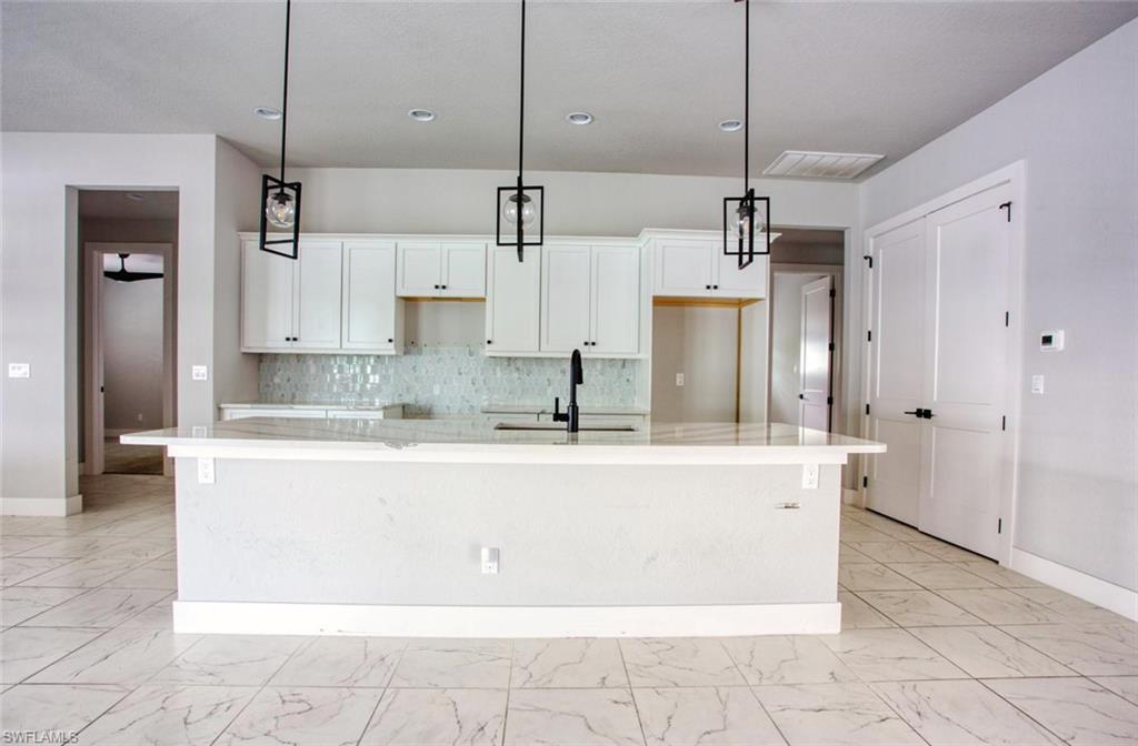 Spacious kitchen featuring an expansive island with a light-toned countertop, white cabinetry with dark hardware, a decorative tile backsplash, and three pendant light fixtures