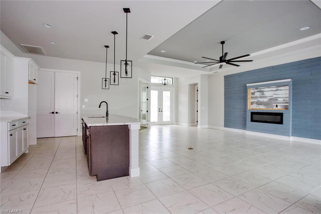 Spacious interior featuring large format tile flooring, a kitchen island with sink, white shaker cabinetry, a ceiling fan, and an accent wall with an inset electric fireplace