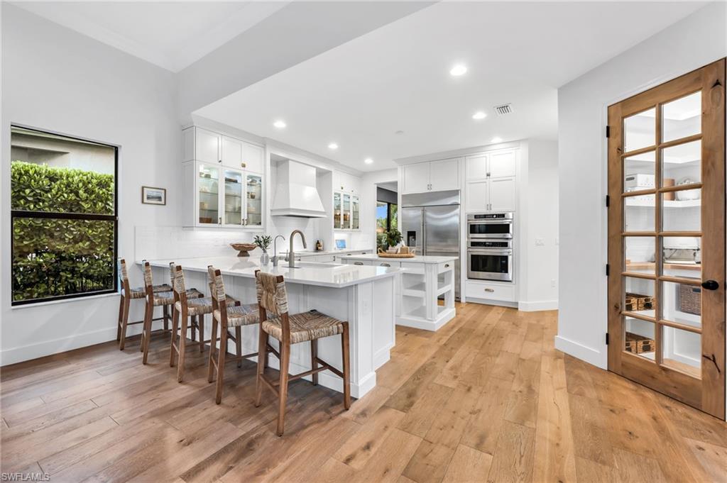 Bright kitchen featuring wood-finish flooring, white cabinetry, a built-in double oven, stainless steel refrigerator, and a pantry with a wood-framed glass door