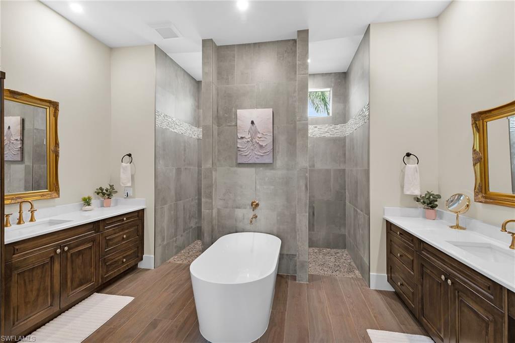 Freestanding soaking tub with brushed gold fixtures, flanked by dual wood-finish vanities featuring white countertops and undermount sinks