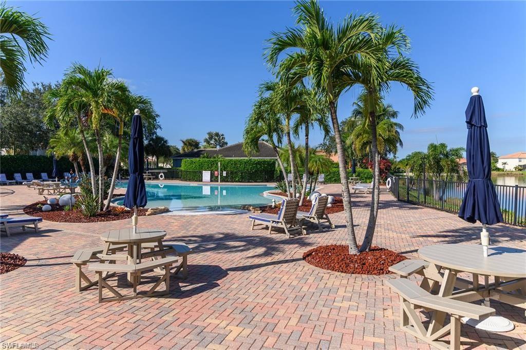 Outdoor pool area featuring a brick paver deck, multiple palm trees, and picnic tables with umbrellas