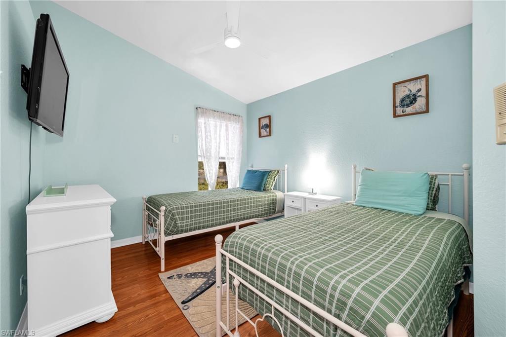 Vaulted ceiling bedroom featuring wood-finish flooring, a window with sheer curtains, a wall-mounted television, and a ceiling fan with integrated lighting