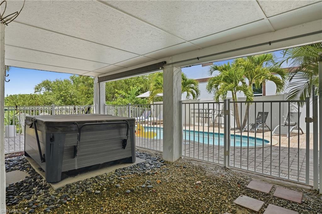 Covered patio featuring a hot tub on a pebble base, opening to a fenced pool area with palm trees and a paved deck - Virtually Edited Image