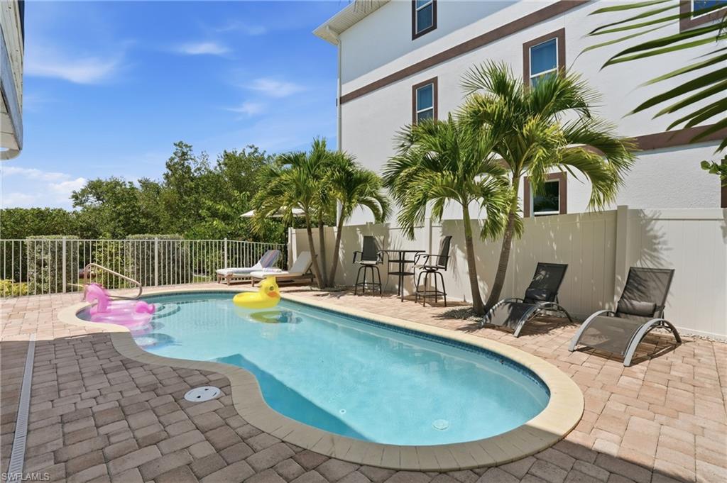 Curved swimming pool with an integrated baja shelf, surrounded by paver decking, a white privacy fence, and mature palm trees - Virtually Edited Image