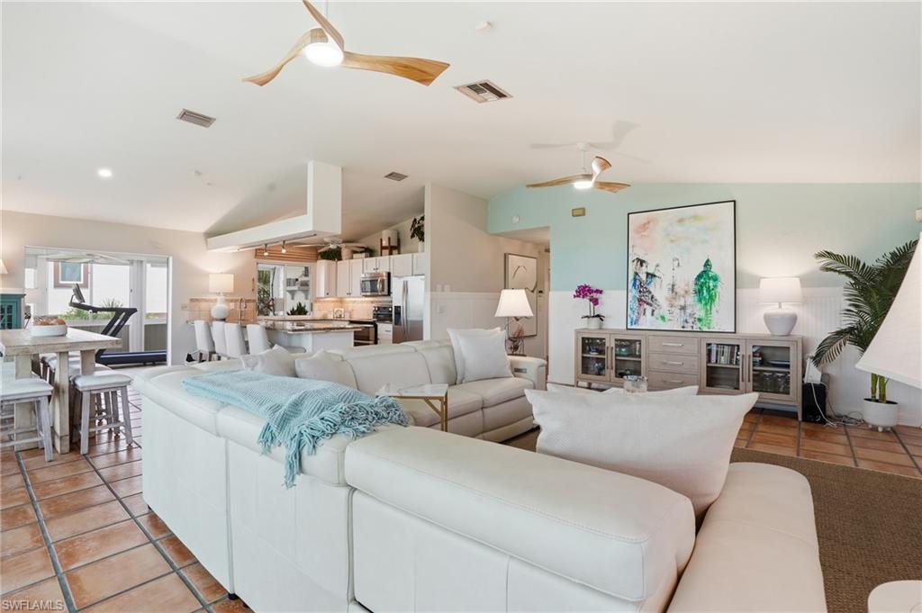 Spacious open-concept living area featuring terracotta tile flooring, vaulted ceilings with modern ceiling fans, a kitchen with white cabinetry and stainless steel appliances, and a breakfast bar with seating - Virtually Edited Image