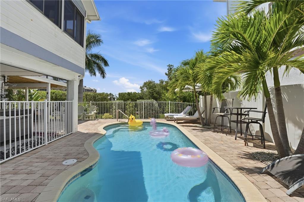Curved swimming pool featuring a paver deck, white perimeter fencing, mature palm trees, and an upper-level screened balcony - Virtually Edited Image