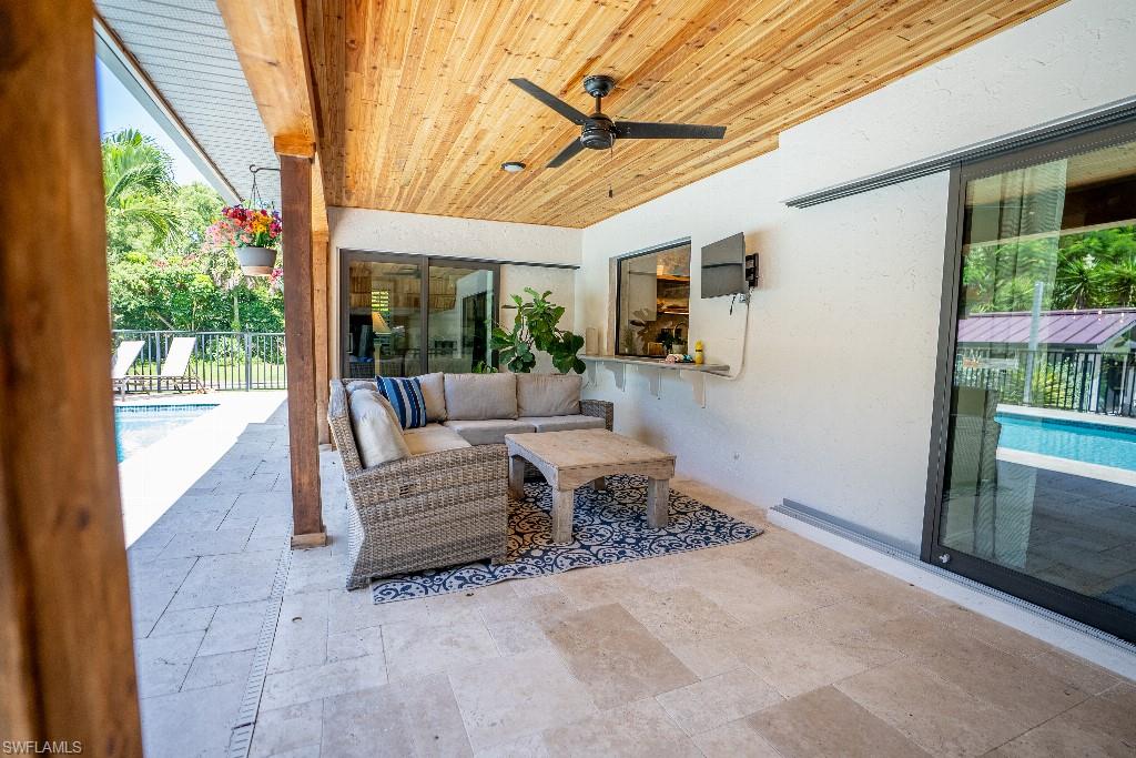 Covered outdoor patio featuring a wood-panel ceiling with a ceiling fan, light-toned stone tile flooring, and a pass-through window with an exterior bar counter
