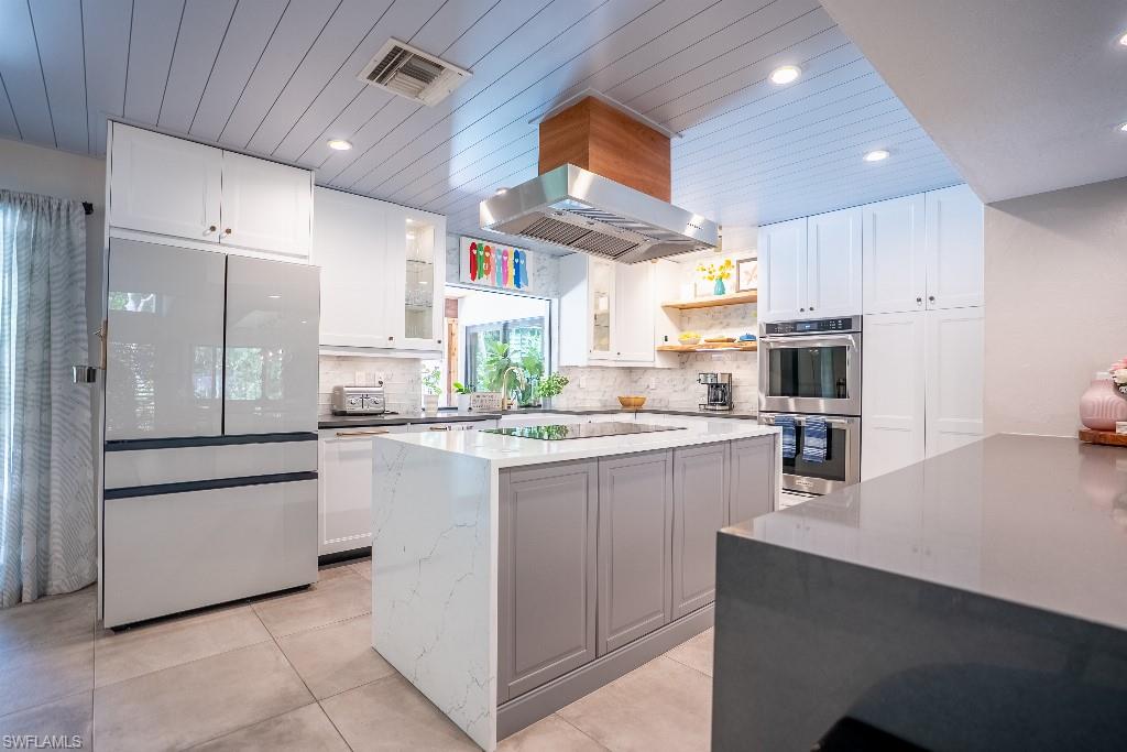Kitchen featuring a center island with waterfall countertop, stainless steel appliances including a built-in oven and range hood, ample white cabinetry, and wood-finish ceiling panels