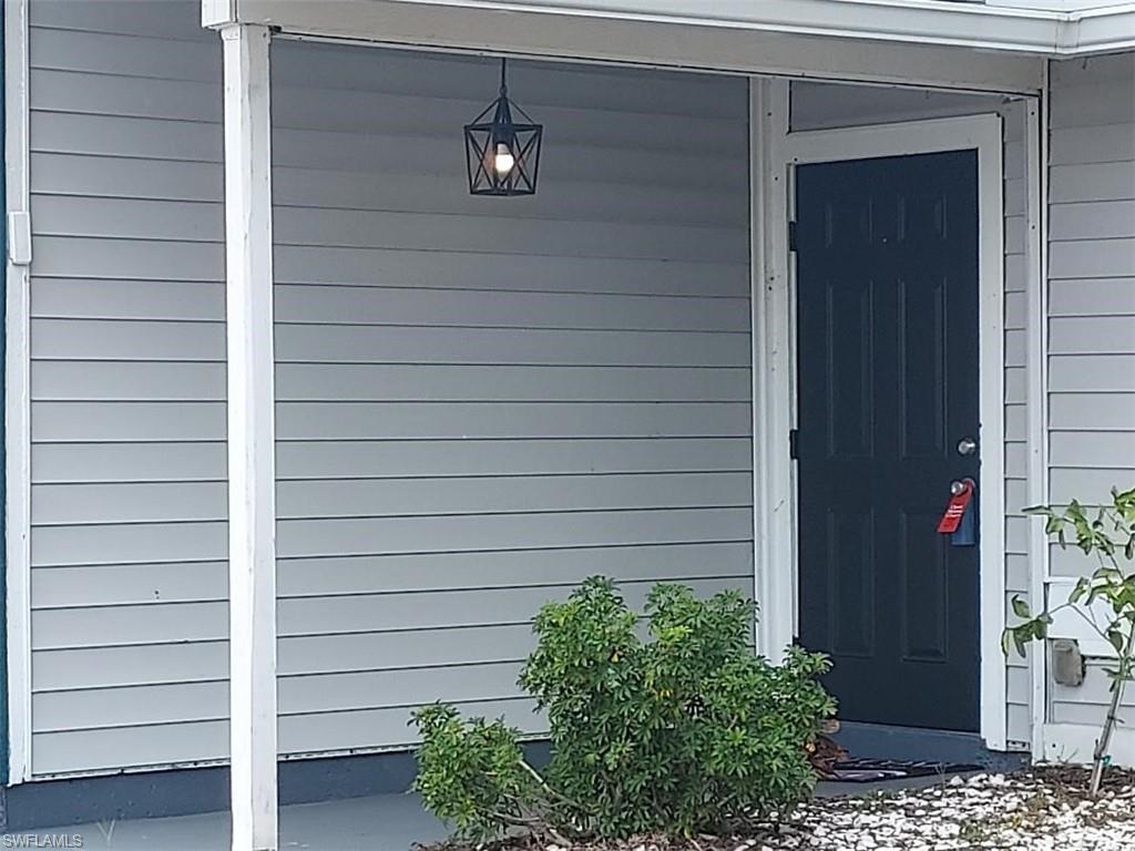 Entryway featuring a dark paneled door, light gray siding, an overhead geometric pendant light, a concrete porch, and established shrubbery