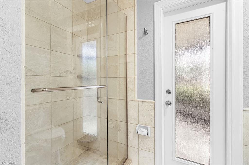 Glass-enclosed shower featuring light-toned square tile, a built-in corner shelf, and a brushed metal handle