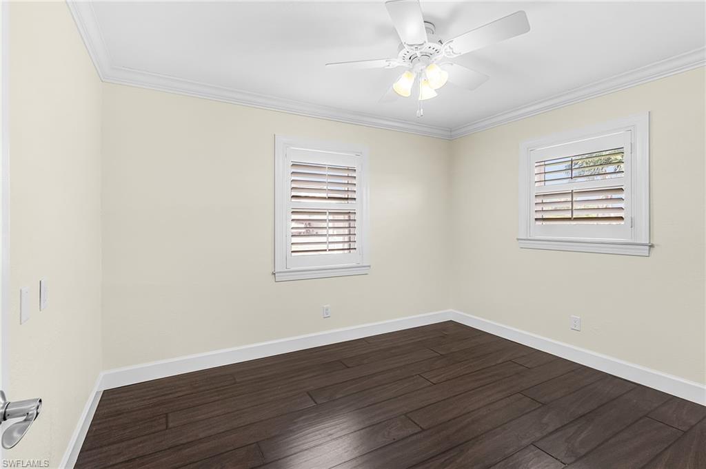 Room featuring dark wood-finish flooring, two windows with white plantation shutters, crown molding, white baseboards, and a white ceiling fan with integrated lighting