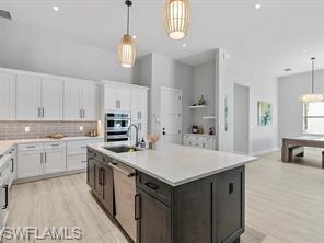 Expansive kitchen featuring a central island with a sink, stainless steel appliances, white cabinetry, a tiled backsplash, and wood-finish flooring