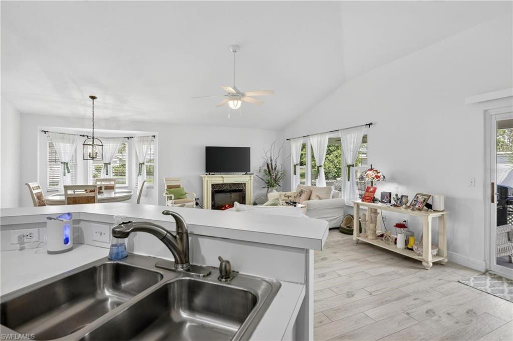 Kitchen bar with a double basin stainless steel sink, extending to a living area featuring vaulted ceilings, light wood-finish flooring, multiple windows, and a sliding glass door