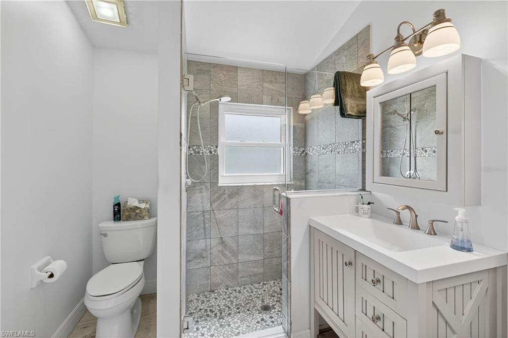 Bathroom featuring a standing shower with clear glass enclosure and pebble floor, gray tile shower surround with decorative mosaic accent, white vanity with integrated sink, brushed nickel faucet, and a wall-mounted medicine cabinet with mirrored door