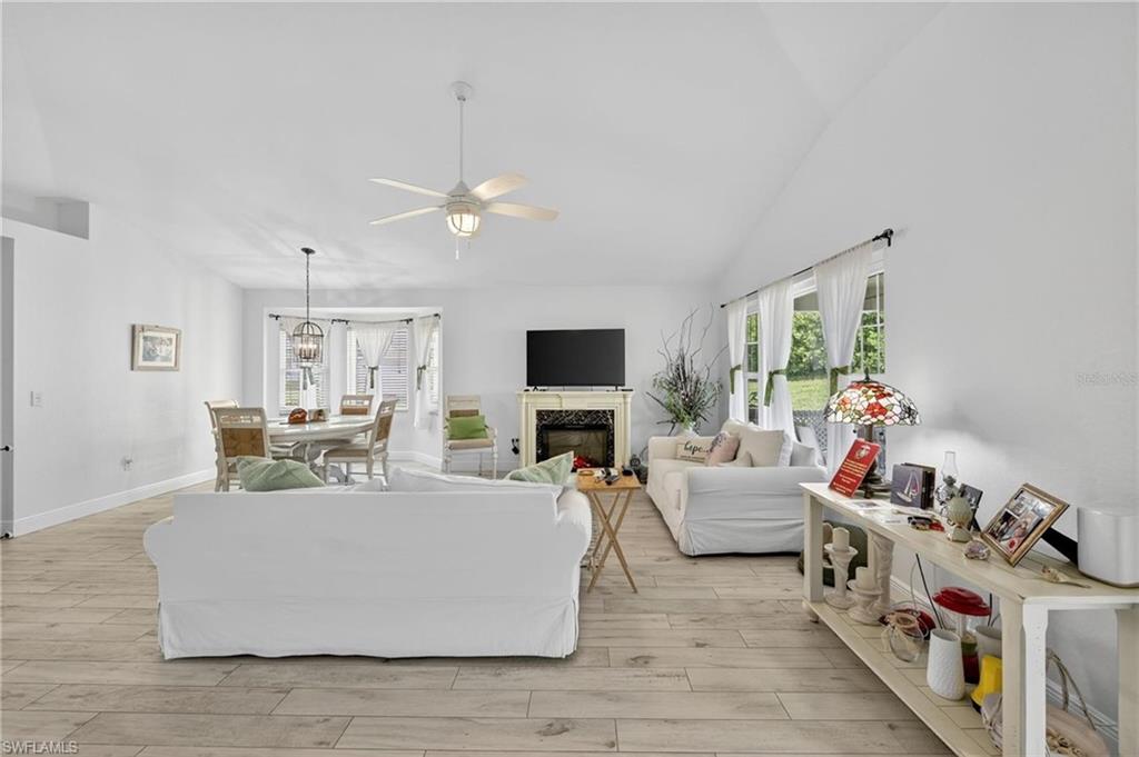 Open-concept living area featuring vaulted ceilings, wood-finish flooring, a ceiling fan with integrated lighting, a fireplace with a mantel, and multiple windows providing natural light