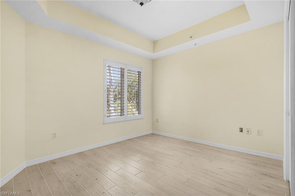 Spacious room featuring light wood-finish flooring, a tray ceiling, and a large window with plantation shutters